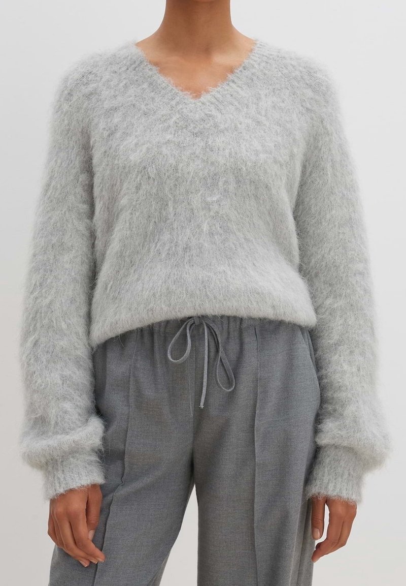 Pull doux gris clair en maille duveteuse à col en V porté avec un pantalon gris taille haute à cordon de serrage, mains détendues le long du corps.