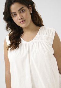 White sleeveless blouse with a V-neck, featuring gathered texture at the shoulders and flowing fabric. Soft and lightweight cotton material.