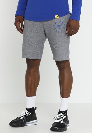 Mann trägt graue Shorts mit dem Schriftzug „All Day Hustle“ und Bull-Logo, blauen Oberteil, weiße Socken und schwarze Sportschuhe mit weißen Schnürsenkeln.