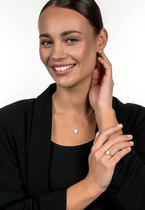 Silver jewelry set featuring a pendant necklace with a circular stone, stud earrings, and a ring with similar stones, against a dark outfit.