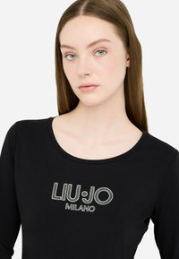 Naisel on sirged pruunid juuksed, seljas must pikka varrukat särk, mille esiküljel on valged studidega "LIU·JO MILANO" logo.