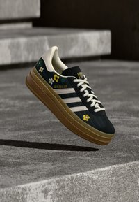 Pantof sport din material textil negru cu dungi albe, broderie florală și talpă de gumă bej. Dispune de guler căptușit și șireturi. Brandul este vizibil pe limbă.