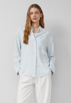 Kobieta o długich blond włosach, ubrana w jasnoniebieską pasiastą koszulę zapinaną na guziki, na białym topie i białych spodniach, stojąca z rękami w kieszeniach.