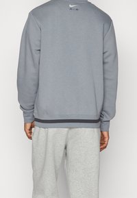 Muž v svetlošedé mikině Nike Air a svetlošedých teplácích, zobrazený zezadu na jednobarevném pozadí.