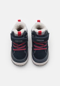 Zapatillas altas de color azul marino con cordones rojos, detalles grises y forro de piel. Presentan una correa de velcro y suela de goma con textura antideslizante.