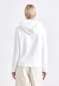 Hvit hettegenser laget av myk stoff, med snøring i hetten, ribbede mansjetter og en avslappet passform. Kombinert med lysebeige bukser.