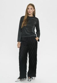 Schwarzes Langarmoberteil mit glitzernder Textur, kombiniert mit schwarzen weiten Hosen, die vertikale Fransenverzierungen und eine glatte Oberfläche aufweisen.