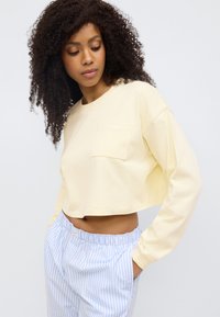 Κροπ μακρυμάνικο πουκάμισο σε ανοιχτό κίτρινο χρώμα με τσέπη στο στήθος, κατασκευασμένο από απαλό βαμβακερό ύφασμα. Συνδυάζεται με παντελόνι με ελαστική μέση σε ανοιχτό μπλε χρώμα με ρίγες.