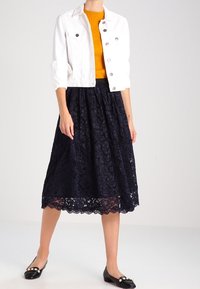 Veste en denim blanc à manches courtes et boutons argentés, associée à une jupe en dentelle bleu marine foncé, avec un ourlet festonné. Ballerines noires avec accents en perles.