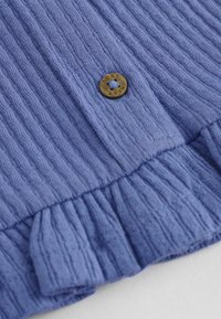 Tissu bleu texturé avec un petit bouton marron gravé du mot « NEXT », présentant des plis cousus le long du bord.
