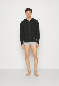 Fekete cipzáras kapucnis pulóver elülső zsebbel, világosszürke fehérneműre húzva. Az anyag puhanak és sima textúrájúnek tűnik. Minimális márkajelzés látható.