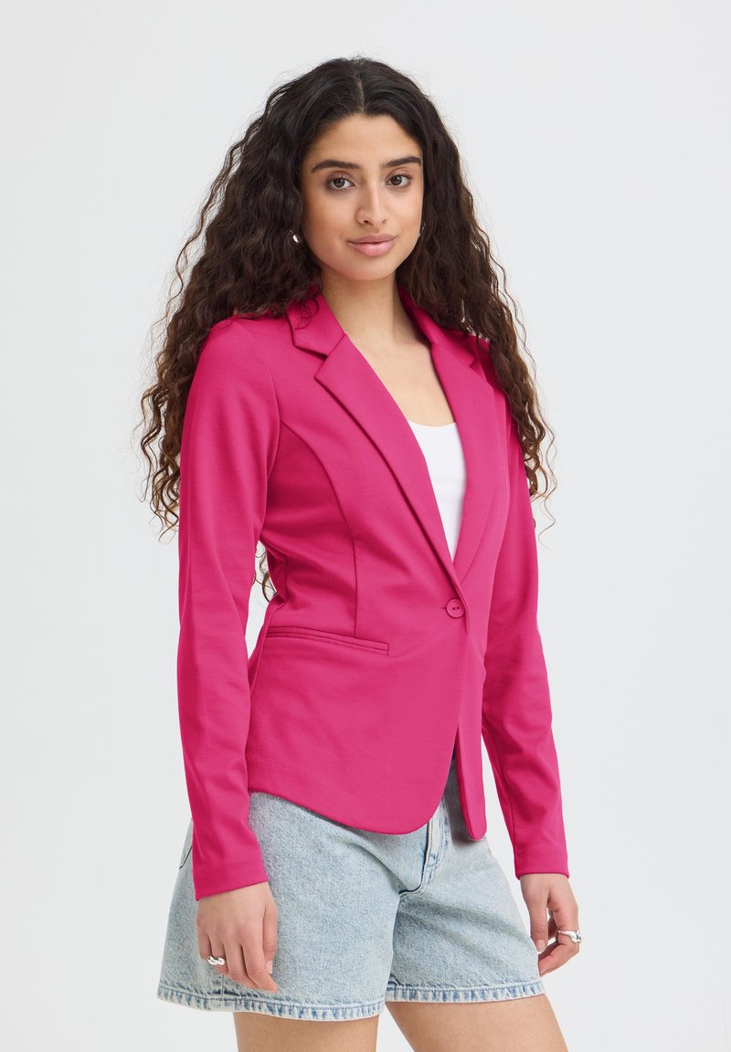 Jeune femme aux longs cheveux bouclés porte un blazer rose vif sur un top blanc et un short en jean clair, debout devant un fond uni.
