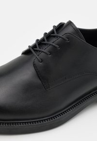Scarpa in pelle nera con texture liscia, punta rotonda e design con lacci. Presenta cuciture sottili e una suola in gomma nera resistente.