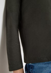 Dunkelgrünes Langarmhemd aus weichem, strukturiertem Material, kombiniert mit hellblauen Jeans aus Denim. Sichtbare Nähte und Saumdetails.