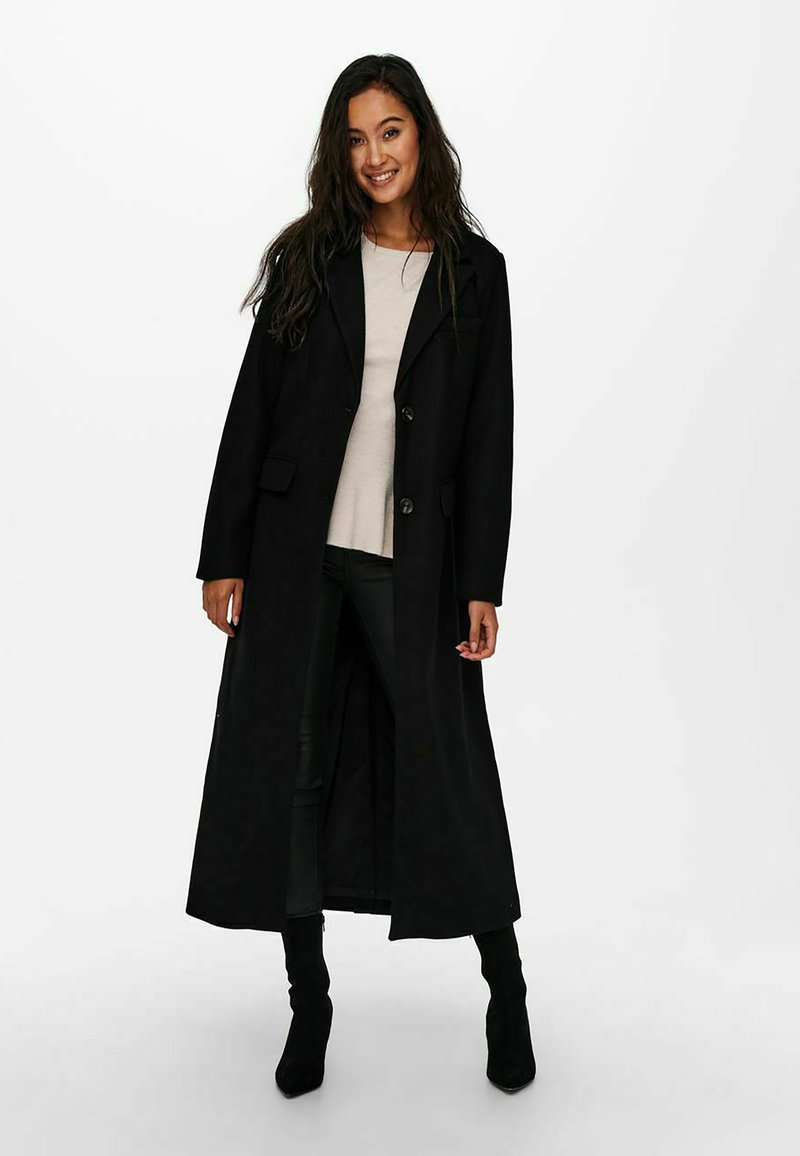Black long coat with a notched collar, front buttons, and side pockets. Worn over a light-colored top and fitted black pants.