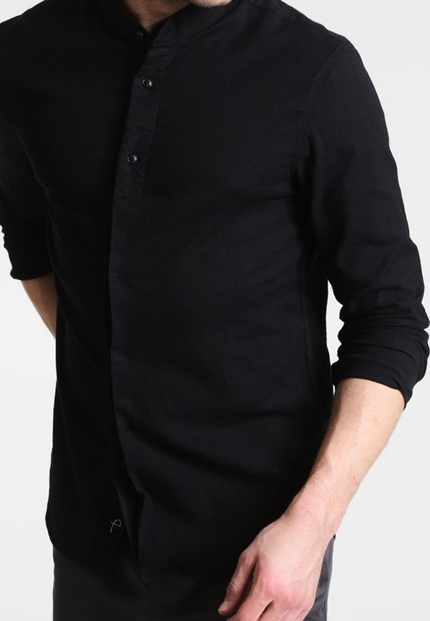 Uomo che indossa una camicia nera a maniche lunghe con i polsini arrotolati, in piedi su uno sfondo bianco semplice.