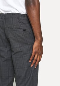 Pantalons à carreaux gris avec une coupe décontractée, dotés d'une poche arrière et d'une étiquette en cuir noir avec le logo EDC. Le tissu semble lisse avec une texture subtile.