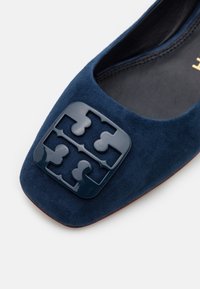 Chaussure plate en daim bleu marine avec un bout carré et une boucle décorative brillante bleu foncé à l'avant.
