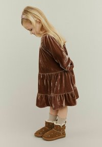 Brun fløjlskjole med lagdelt kant, lange ærmer; parret med hyggelige lysebrune støvler og strikkede cremefarvede sokker, hår prydet med et glitrende pandebånd.