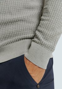 Lichtgrijze gebreide trui met een textuur en ribbels aan de mouwen, met een subtiel patroon. Gecombineerd met marineblauwe broek, hand in de zak.