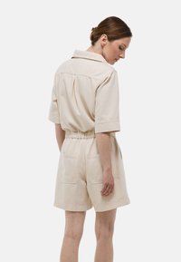 Rompers beige courts avec col, manches courtes, taille élastique et deux poches arrière. Fabriqué dans un tissu doux et durable.
