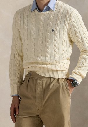 Man wearing cream cable-knit sweater over blue collared shirt, beige pleated trousers, and a wristwatch with brown leather strap.