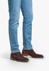 Homme portant un jean bleu clair et des chaussures habillées en cuir texturé marron foncé, se tenant sur un fond blanc.