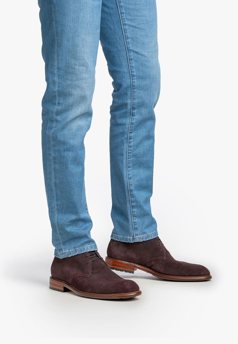 Homme portant un jean bleu clair et des chaussures habillées en cuir texturé marron foncé, se tenant sur un fond blanc.