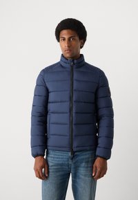 Veste matelassée bleu marine avec un col haut, design matelassé horizontal et fermeture éclair, dotée de poignets élastiques et d'un logo discret sur la manche.