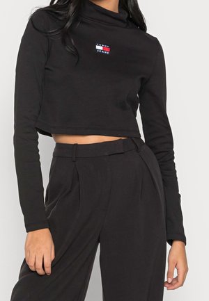 Kobieta w czarnym, krótkim topie z długim rękawem z logo Tommy Jeans oraz czarnych, wysokich plisowanych spodniach na jednolitym tle.