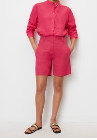 Camisa de lino rosa con cuello alzado y cierre de botones, combinada con shorts de cintura alta a juego y sandalias planas con una correa decorativa.