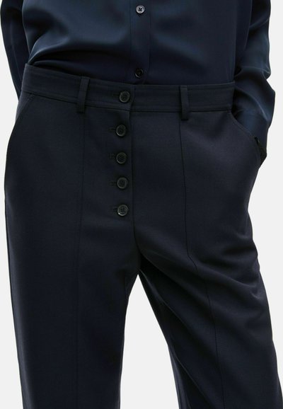 Nahaufnahme einer Person, die dunkelblaue Hosen mit einer Fünf-Knopf-Verschluss und einem passenden dunkelblauen Hemd trägt, mit den Händen in den Taschen.