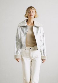 Chaqueta plateada metálica cortada con un gran cuello y puños de borrego, combinada con una blusa beige y pantalones blancos de talle alto.