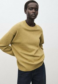 Maglione giallo senape con collo rotondo e polsi a coste, abbinato a jeans scuri. Texture morbida, vestibilità comoda, design semplice.