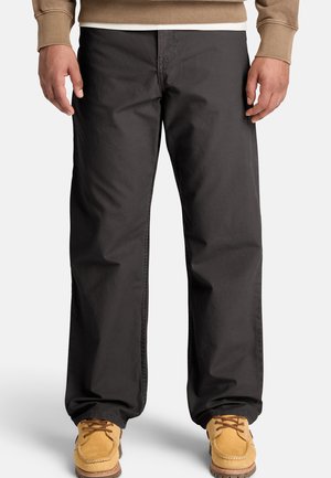 Hombre que lleva pantalones negros holgados y botas marrón claro con cordones, de pie con las manos relajadas a los lados sobre un fondo blanco liso.