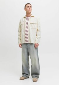 Man in een crèmeskleurige button-up jacket over een gestreept shirt, met losse vervaagde jeans en beige casual schoenen, staande tegen een effen achtergrond.