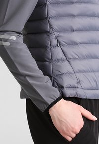 Veste isolante grise avec une texture matelassée, dotée d'une manche grise lisse et extensible, ainsi que d'une petite poche zippée sur le côté.