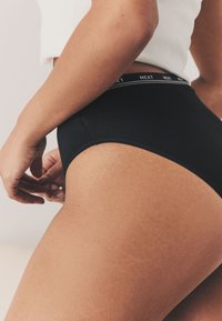 Bragas negras de algodón de cintura alta con textura suave y una delgada cintura elástica con el logotipo de la marca repetido. Vista en primer plano que muestra el ajuste y estilo.