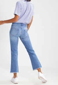 Persona wearing jeans de dobladillo deshilachado de color azul claro, camiseta de rayas de manga corta y zapatillas blancas, de pie con una pierna ligeramente doblada.