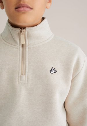 Gai beige puskrekls ar pusi rāvējslēdzēju, izgatavots no mīkstas auduma, ar teksturētu virsmu un mazu melnbaltu putnu logotipu uz krūšu daļas.