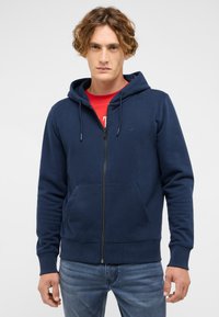 Marineblauer Kapuzenpullover mit Reißverschluss aus Baumwollmischung, mit Fronttasche, Kordelzugkapuze sowie gerippten Bündchen und Saum. Über einem roten Shirt getragen.