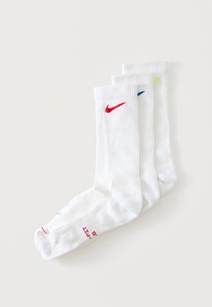 Trzy pary białych skarpetek za kostkę z prążkowanymi ściągaczami, każda z czerwonym, niebieskim i neonowożółtym logo Nike z przodu na górze.
