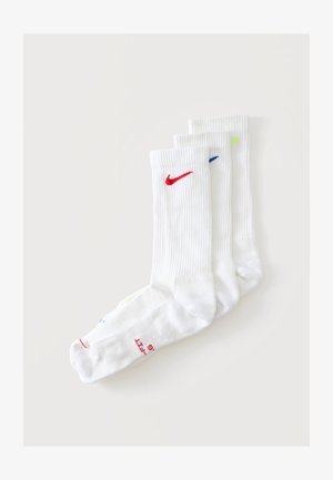 Trois paires de chaussettes blanches montantes avec bord-côtes, chacune présentant un logo Nike en rouge, bleu et jaune fluo sur le dessus devant.