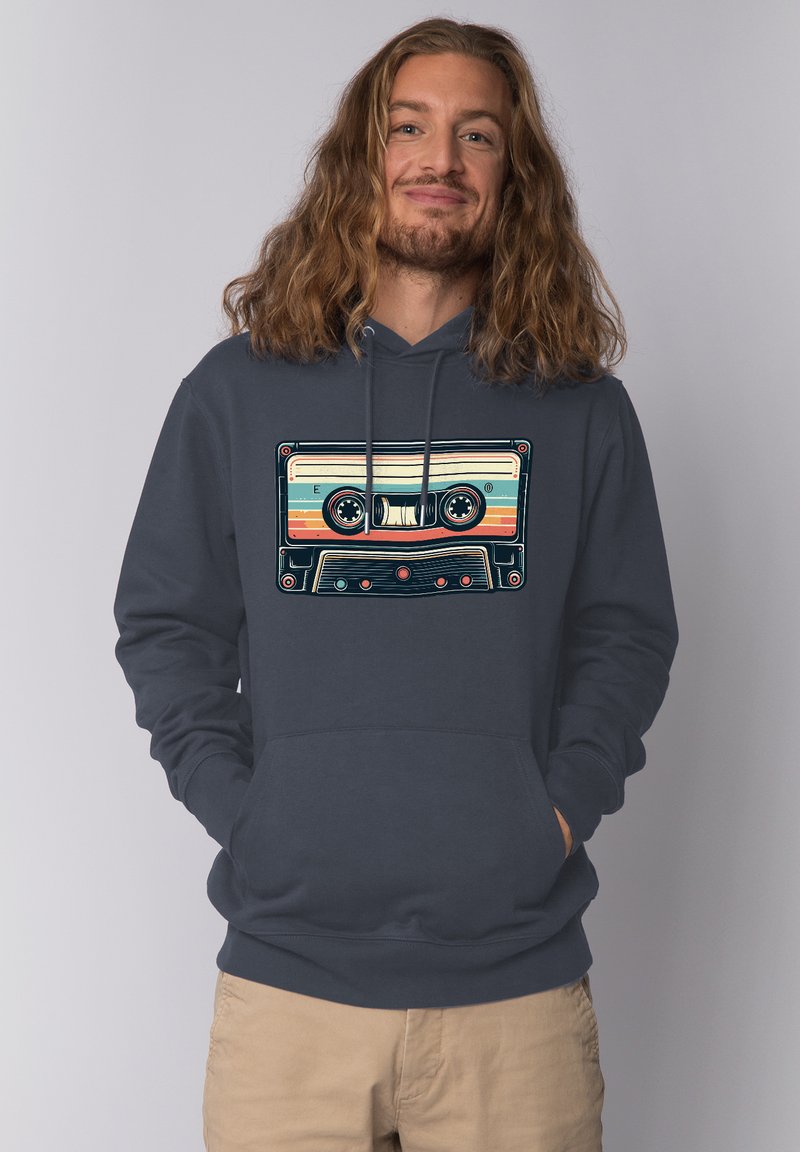 Mann mit langen Haaren und Bart, der einen dunklen Hoodie mit farbenfroher Retro-Kassettenspieler-Grafik trägt, Hände in den Taschen, beige Hose, neutraler Hintergrund.