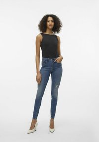 Juodas be rankovių viršus, derinamas su priglundančiais mėlynais džinsais su nusidėvėjimo detalėmis ir baltais aukštakulniais, prieš vienodą foną.