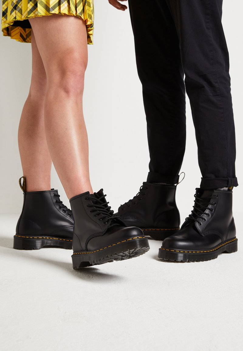 Deux personnes portent des bottes en cuir noir à lacets avec des coutures jaunes, l’une en jupe à carreaux jaune, l’autre en pantalon noir, sur un sol blanc.