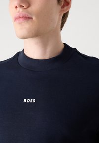 Marineblå hettegenser med ribbestrikket halslinning og hvit "BOSS" logo på brystet. Stoffet ser glatt ut med en tettsittende design.
