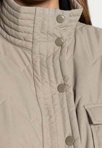 Chaqueta acolchada beige con cuello alto, botones a presión y costuras decorativas. Incluye un bolsillo frontal para mayor utilidad.