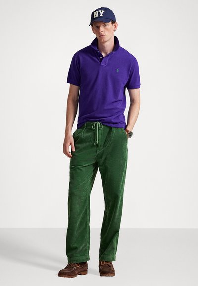 Camisa polo em algodão roxo, calças de veludo verde e sapatos castanhos com atacadores. Acessórios incluem um boné navy com "NY" em branco.