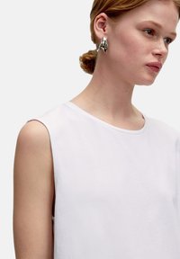Haut sans manches blanc avec encolure ronde ; tissu lisse et léger ; modèle portant une boucle d'oreille en métal distinctif. Design simple, sans motifs.